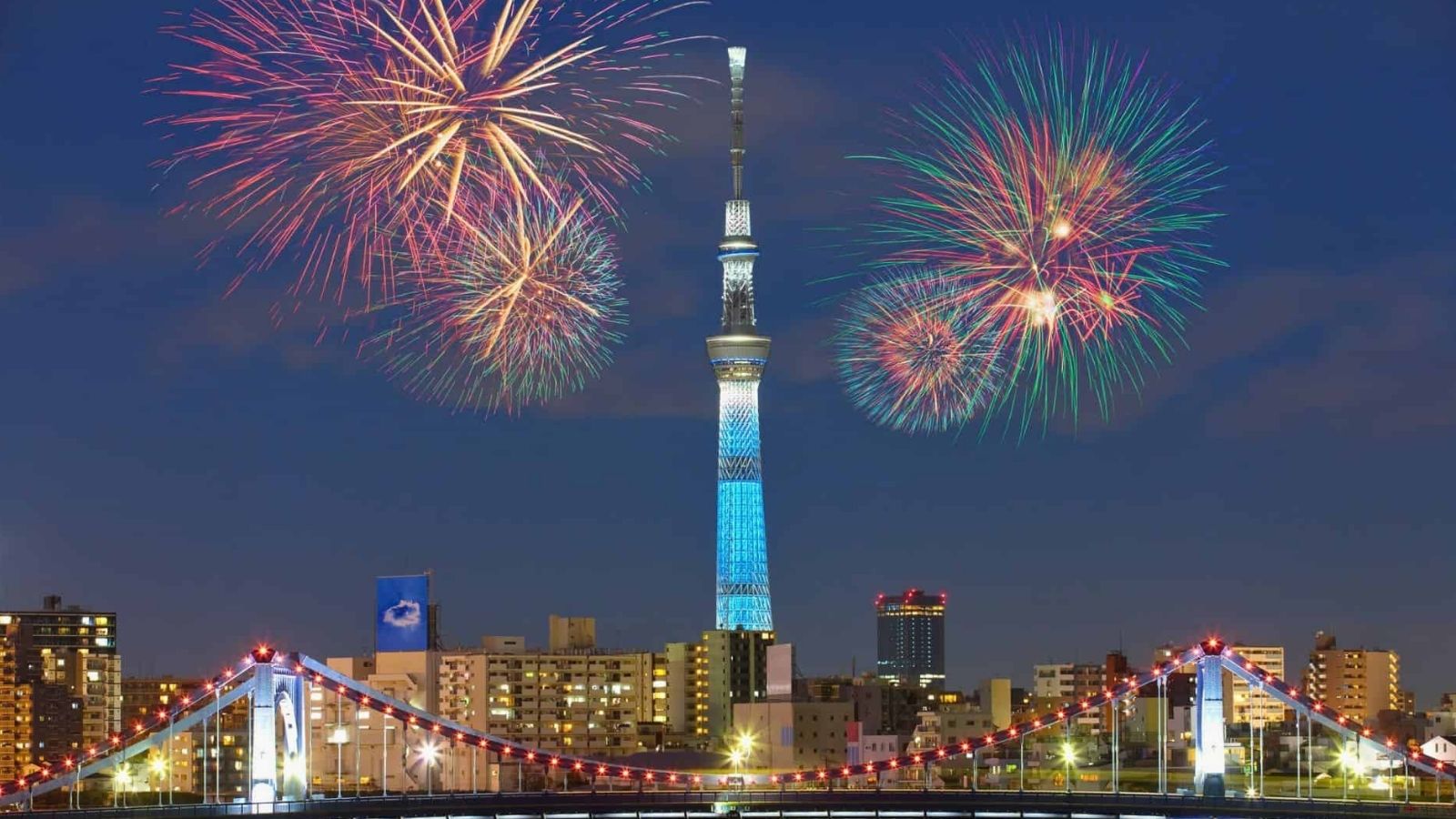 Fireworks light up the night sky above Tokyo Skytree, city buildings, and a glowing bridge in the foreground.