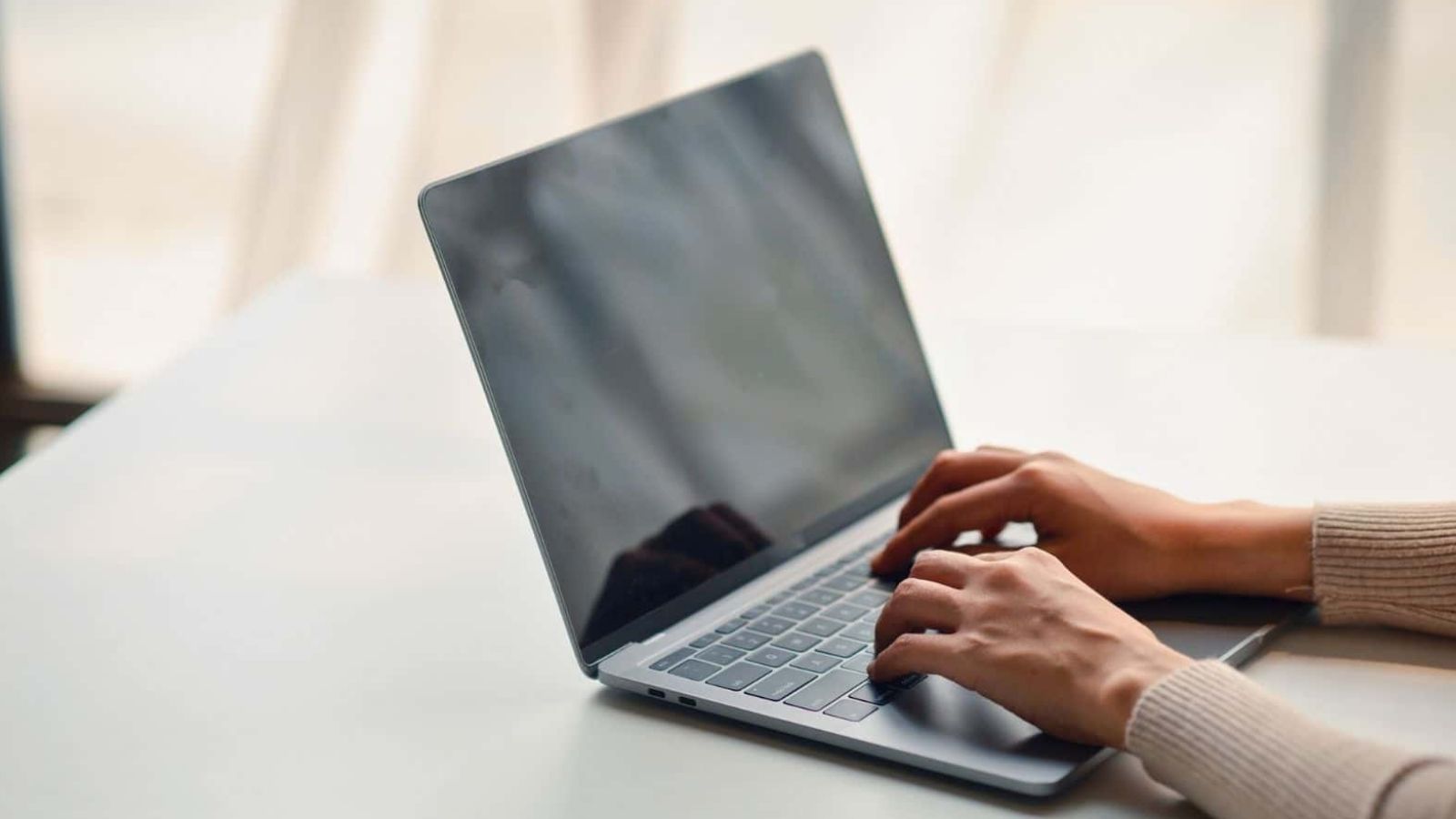 Hands typing on a laptop at a white desk with a light curtain in the background.