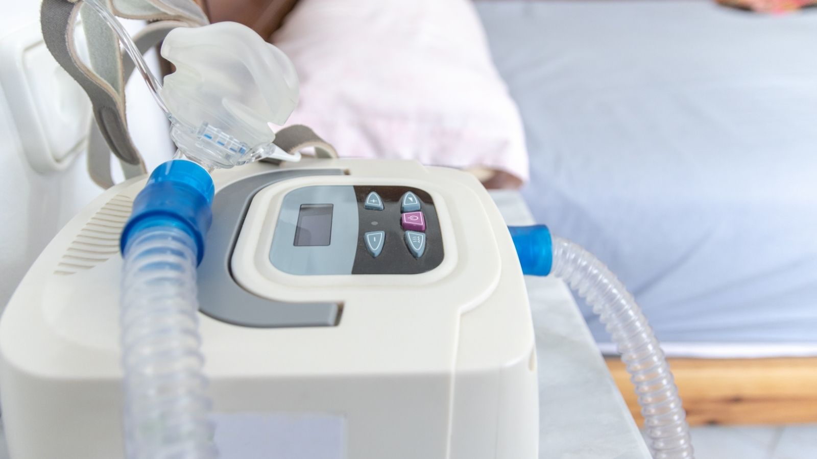 A CPAP machine with mask and tubing rests on a bedside table beside a bed with pillows and sheets.
