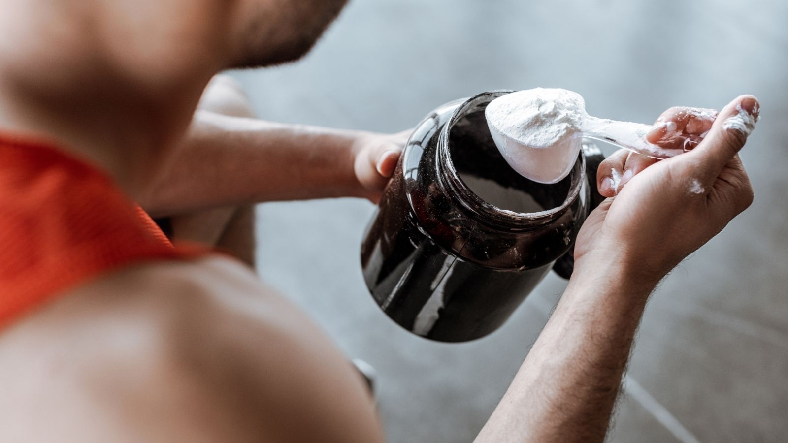 A person in a sleeveless shirt uses a plastic scoop to take white powder from a large black container.