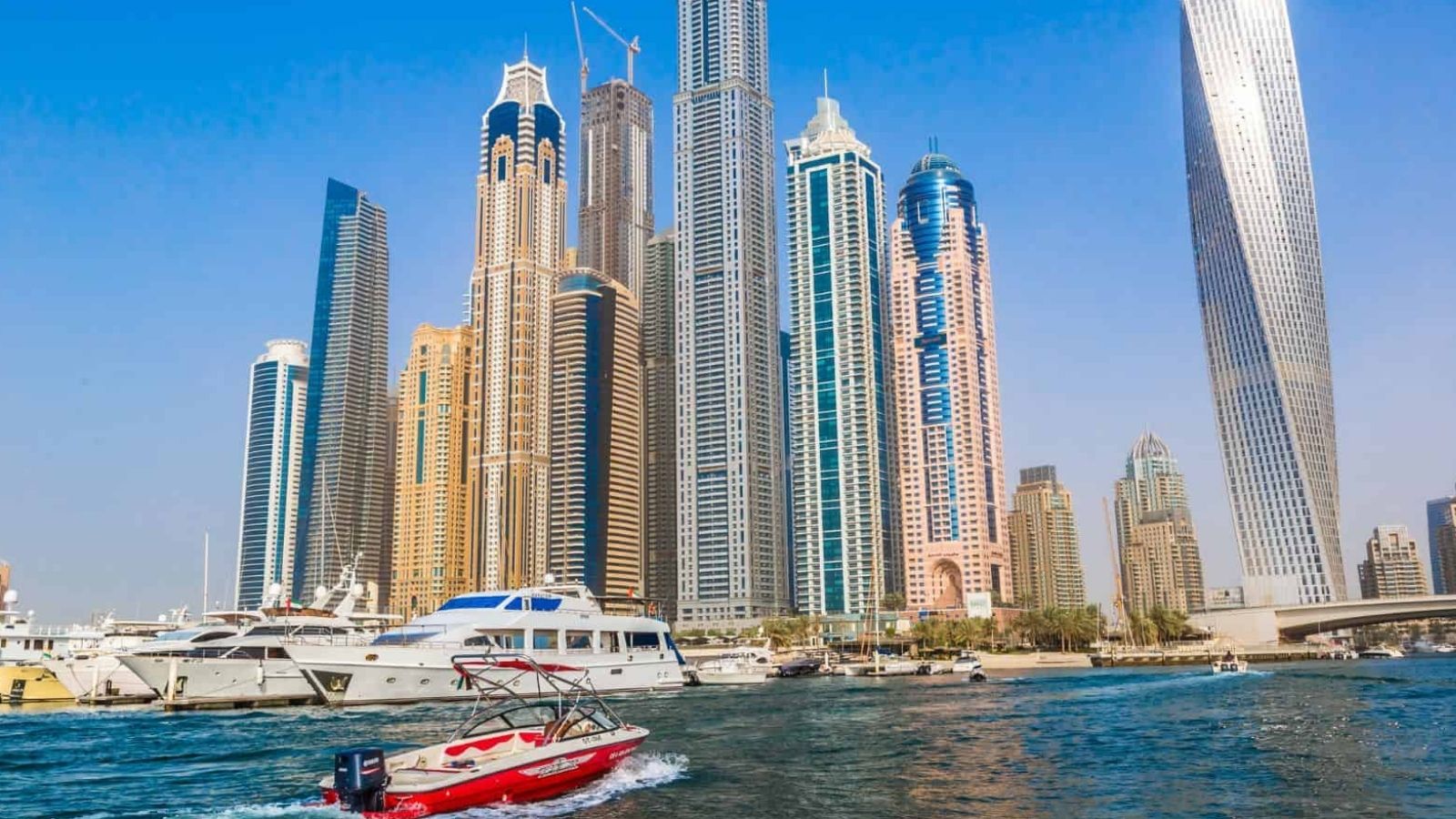 Skyscrapers line the waterfront as yachts and a speedboat cruise in front under a clear blue sky.