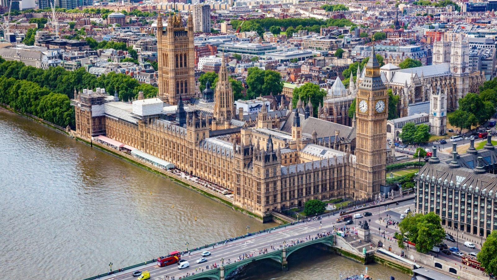 Aerial view of the Houses of Parliament, Big Ben, and Westminster Bridge next to the Thames in London with city buildings nearby.