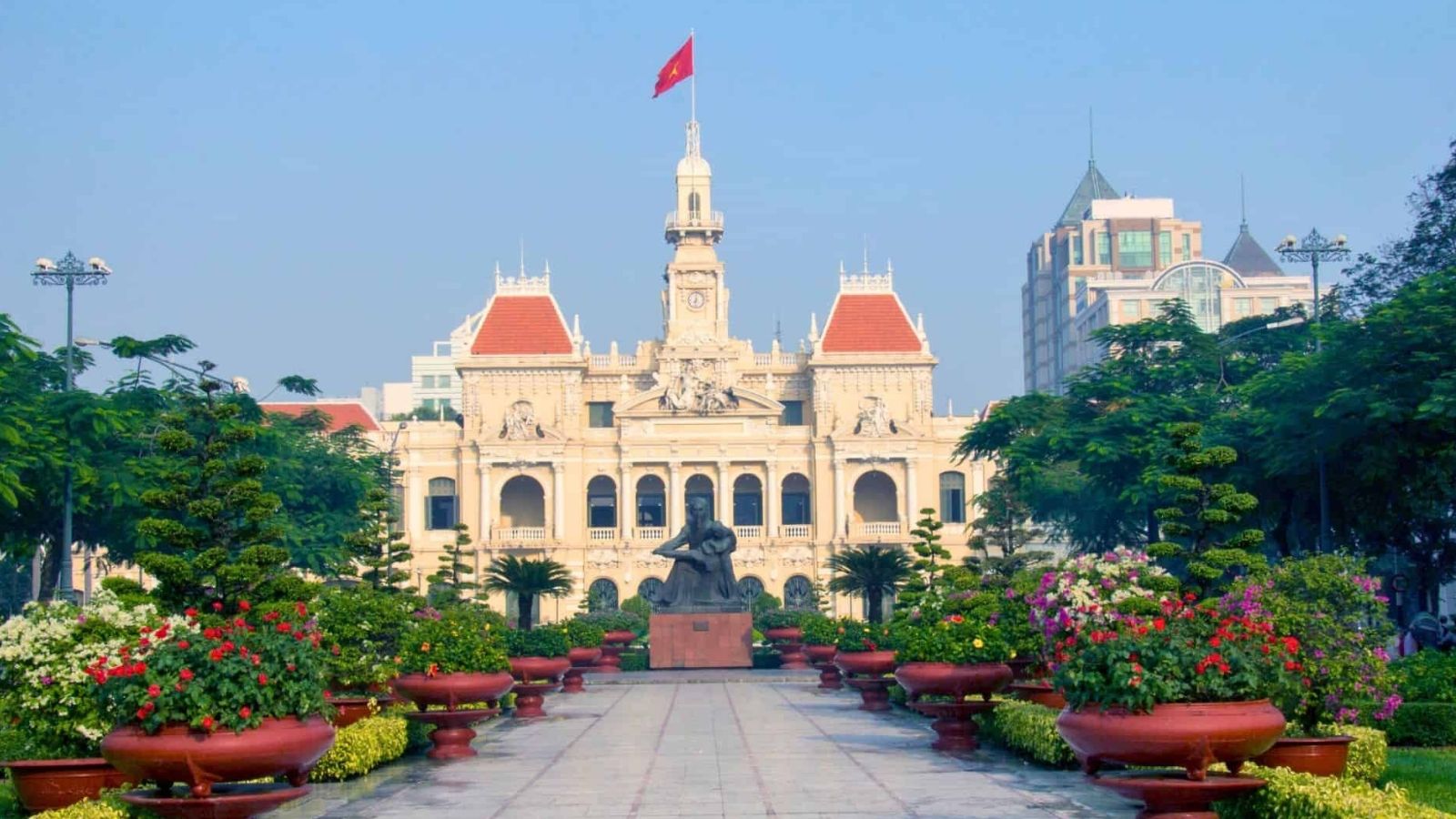 Ho Chi Minh City Hall in Vietnam stands before a potted plant-lined path and statue, beneath a clear blue sky.