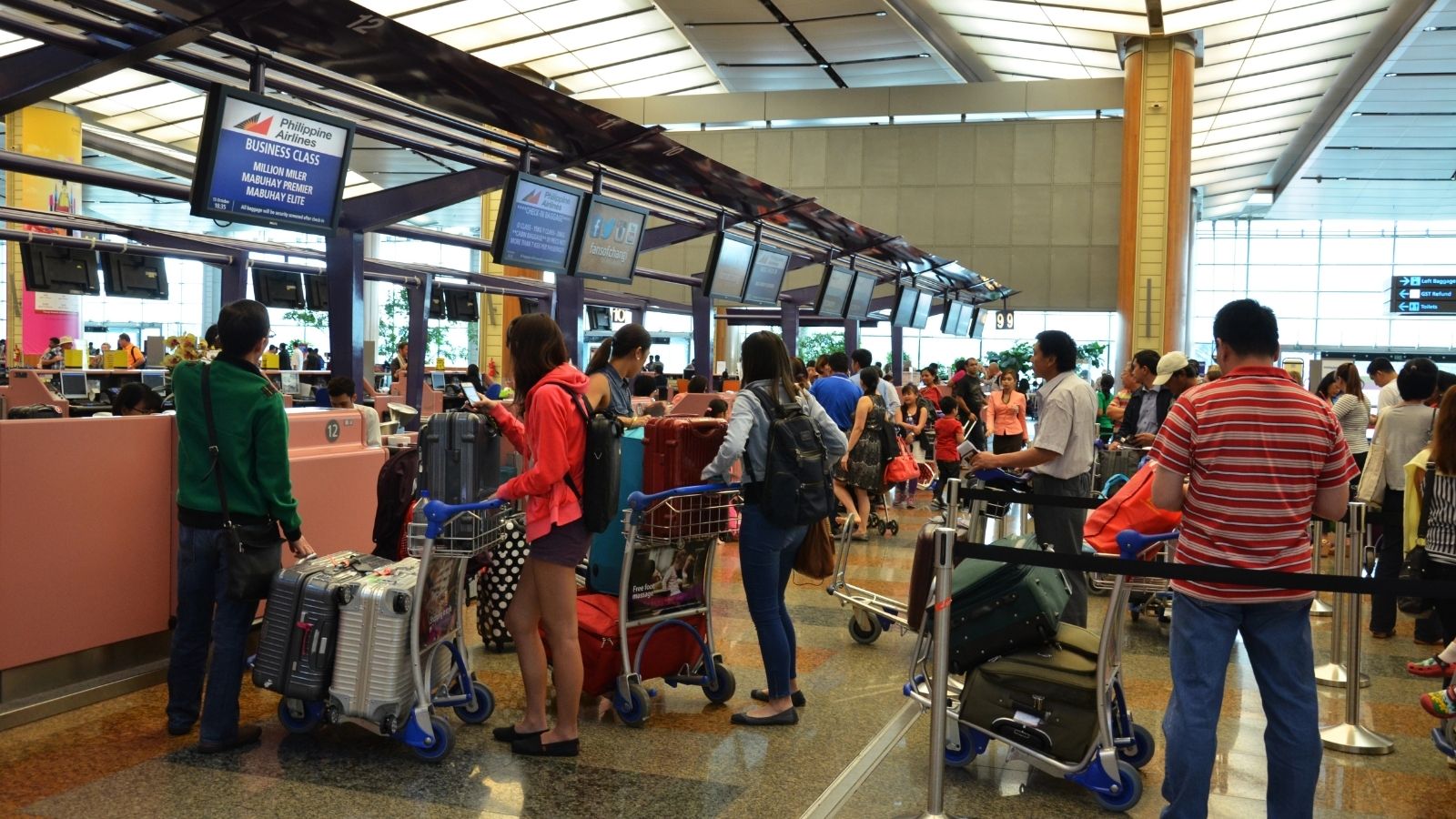 Travelers with luggage carts line up at airport check-in counters beneath overhead monitors in a bright, busy terminal.