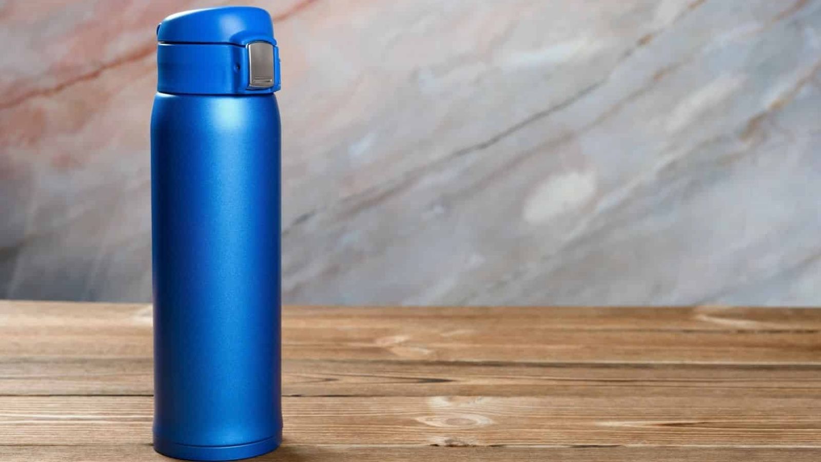 Blue stainless steel water bottle with flip-top lid on wooden surface, set before a marble-patterned background.
