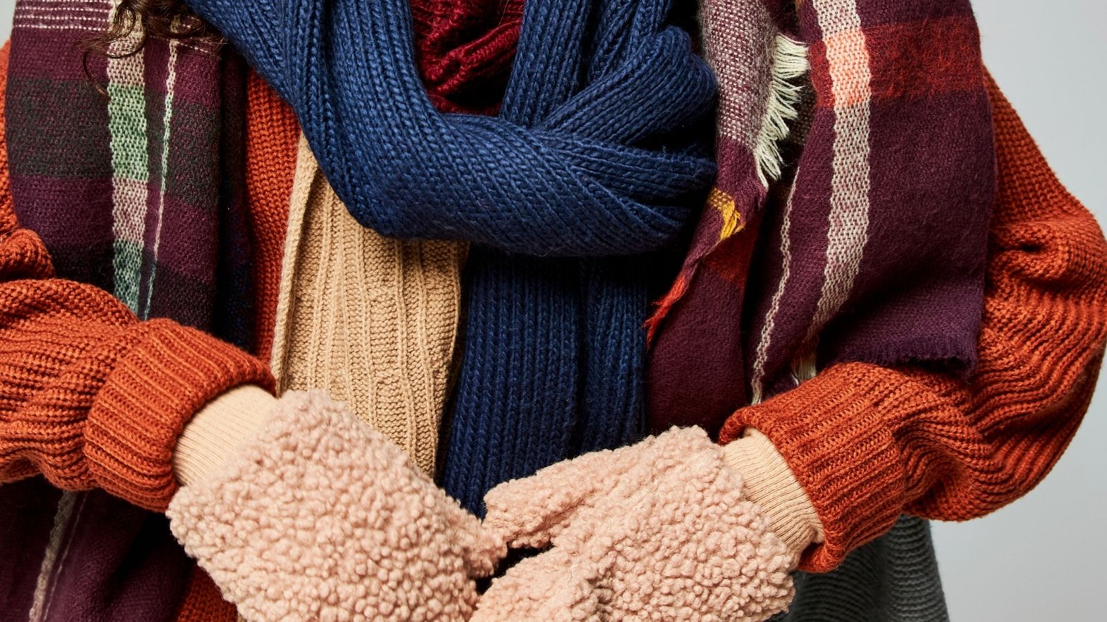 Person in thick knit sweaters, plaid and blue scarves, and beige fleece gloves clasps hands in front.