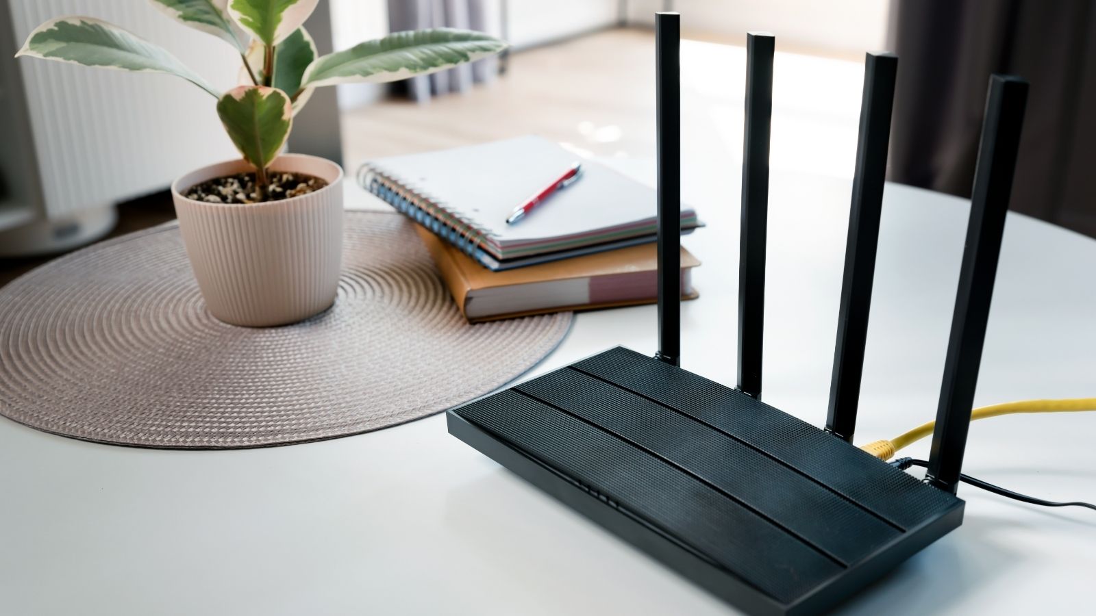 A black wireless router with four antennas is on a white table beside a potted plant, stacked notebooks, and a red pen.