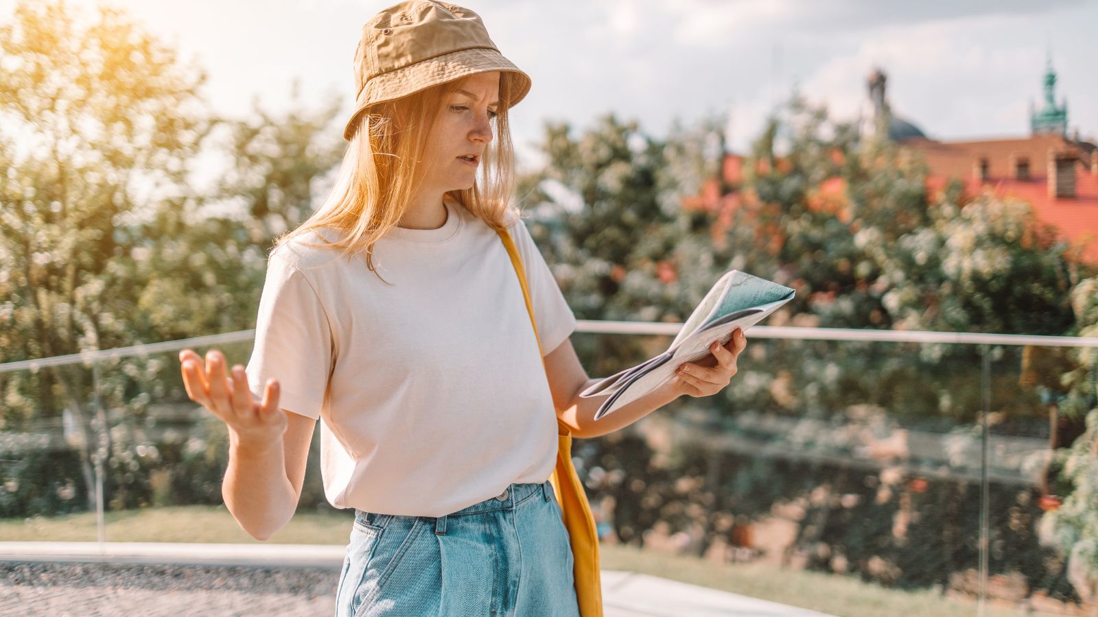 A young woman in a bucket hat and casual clothes looks confused holding a map outdoors with trees and buildings behind her.