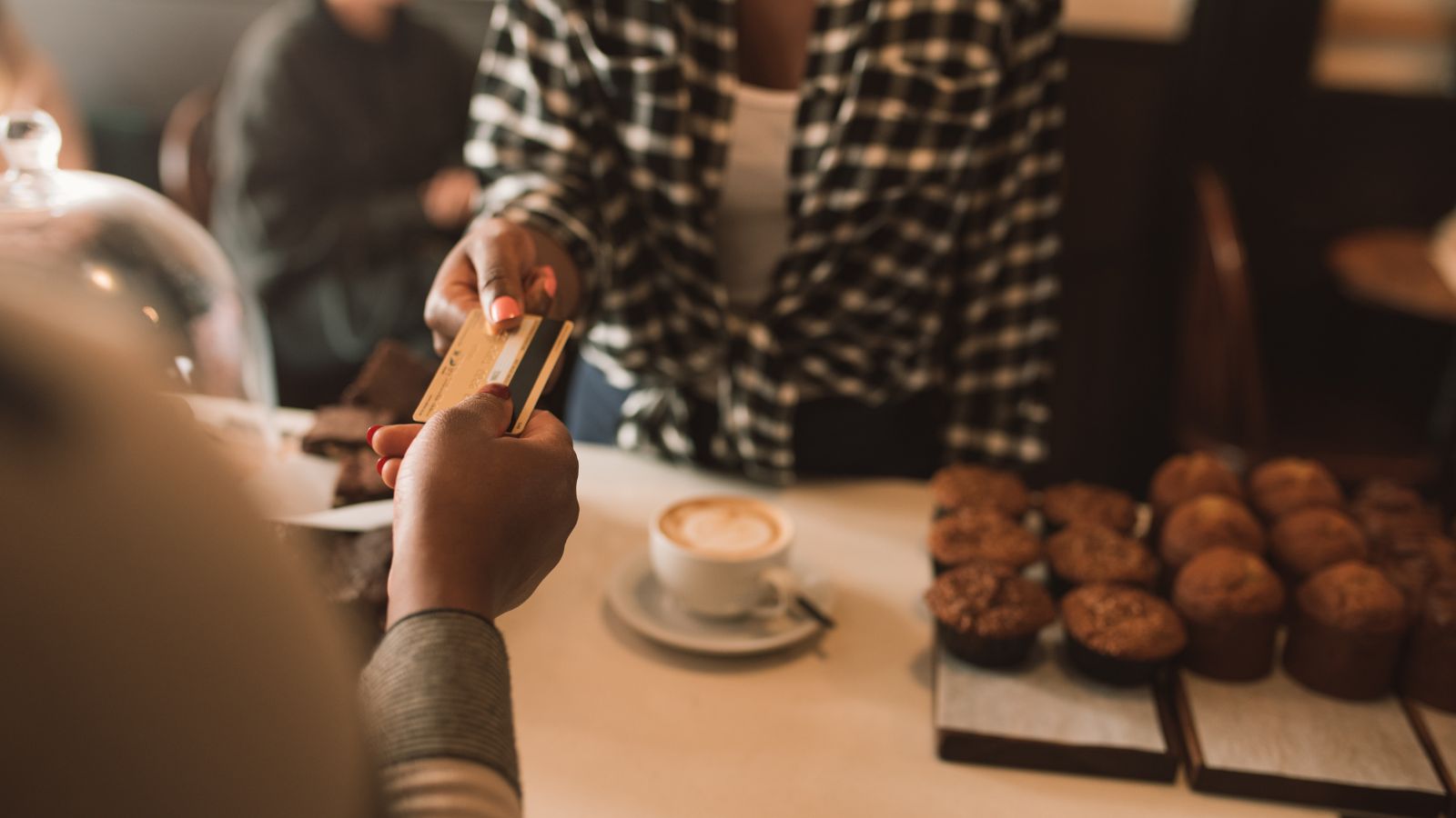 A person gives a credit card to a cafe cashier, with muffins and coffee on the counter.