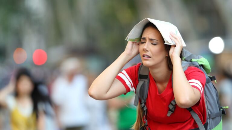 A tired-looking woman in a red shirt shades herself with a map outdoors, with blurred people and trees in the background.
