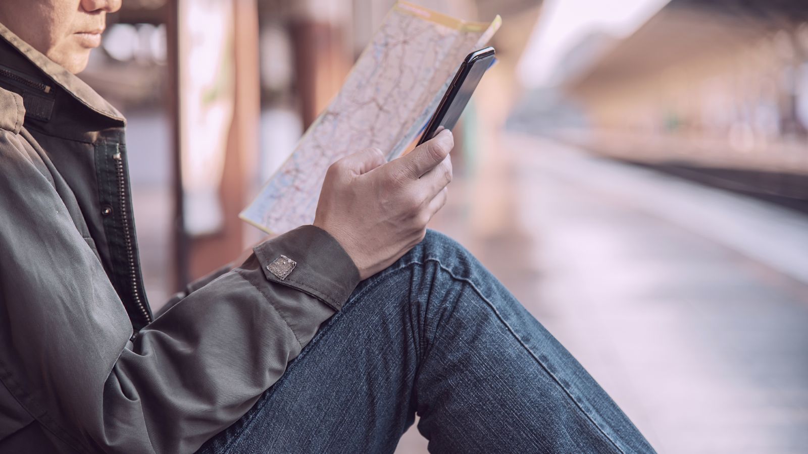A person on a train station bench holds a map in one hand and a smartphone in the other.