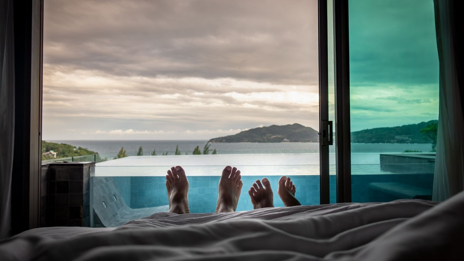 Two people relaxing on a bed with their feet in view, overlooking an infinity pool and a calm ocean with distant hills under a cloudy sky
