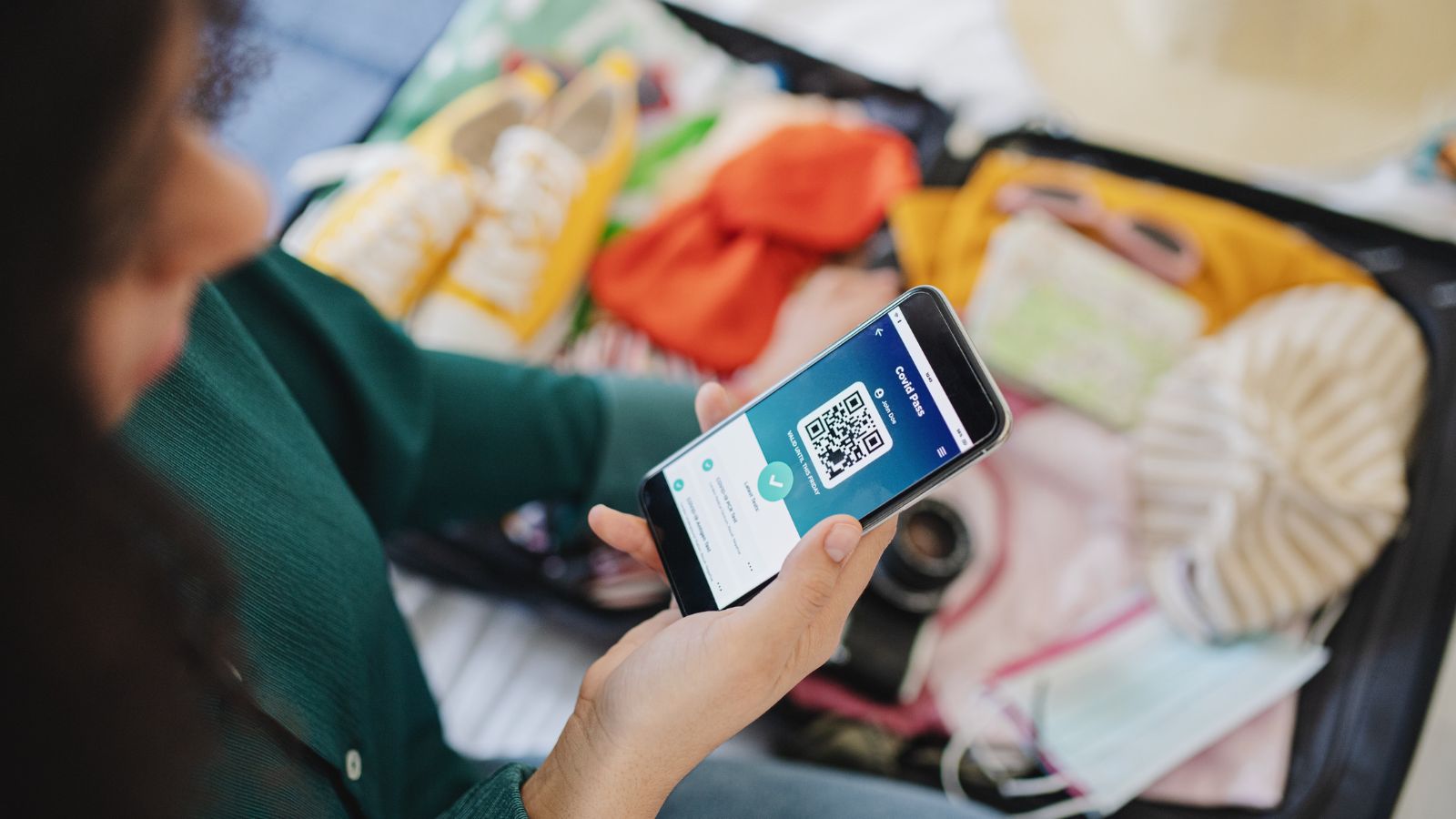 A person packing a suitcase with clothes, shoes, hat, and face mask holds a smartphone displaying a QR code.