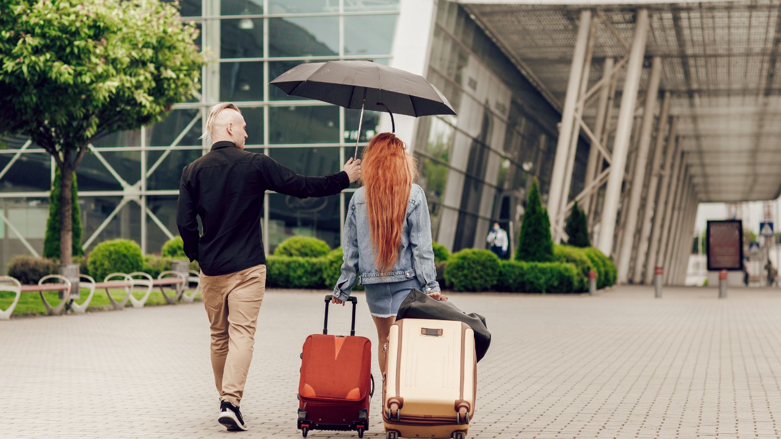 A back photo of a couple where the woman is carrying two luggage and the man is covering her head with the umbrella.