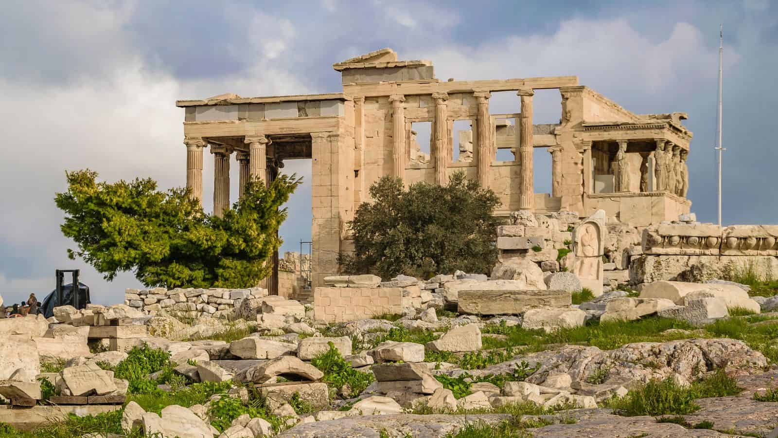 Erechtheion temple ruins with columns and trees on the Acropolis in Athens, Greece, beneath a partly cloudy sky.