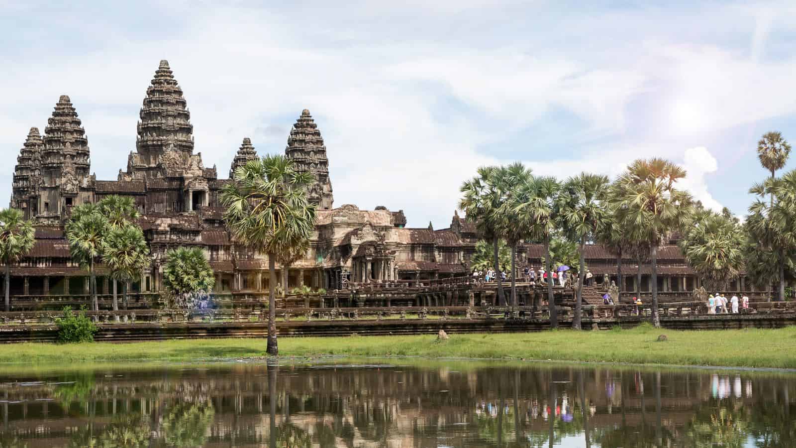 Angkor Wat temple, stone towers, palm trees, and tourists reflected in a pond beneath a partly cloudy sky.