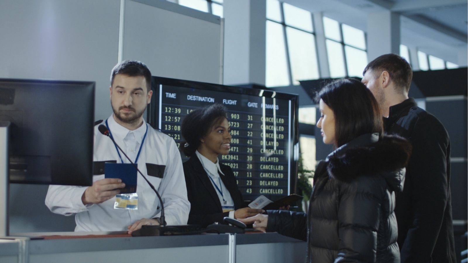 Airline staff assist passengers at an airport check-in counter as a departure board behind them lists several cancelled flights.