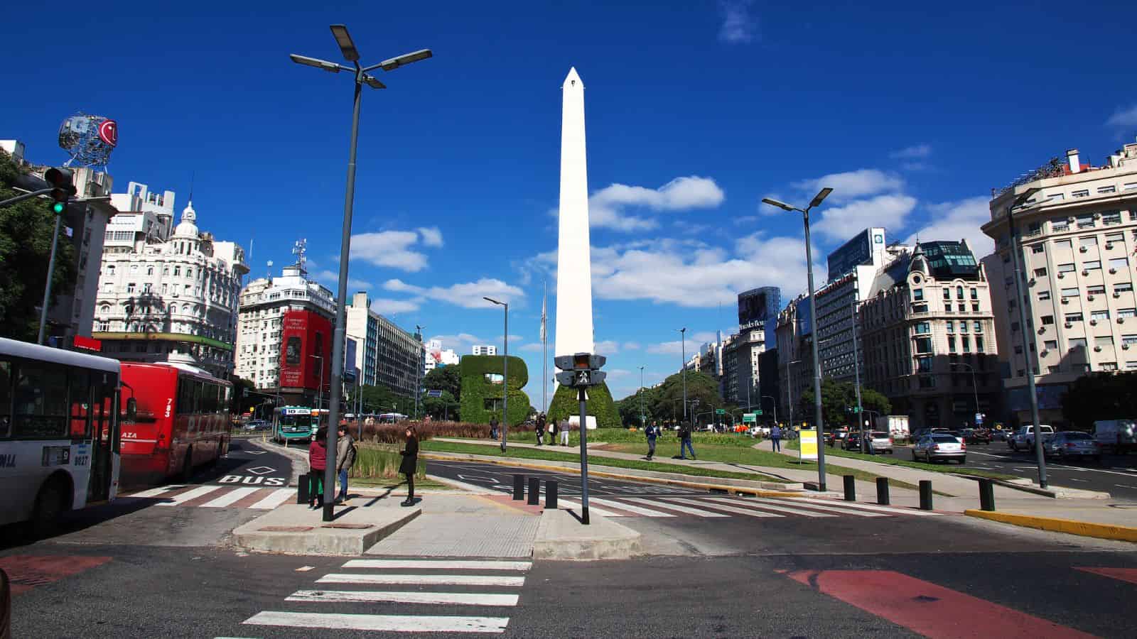 A wide street leads to the white Obelisk of Buenos Aires, amid buildings, buses, and pedestrians under a partly cloudy sky.