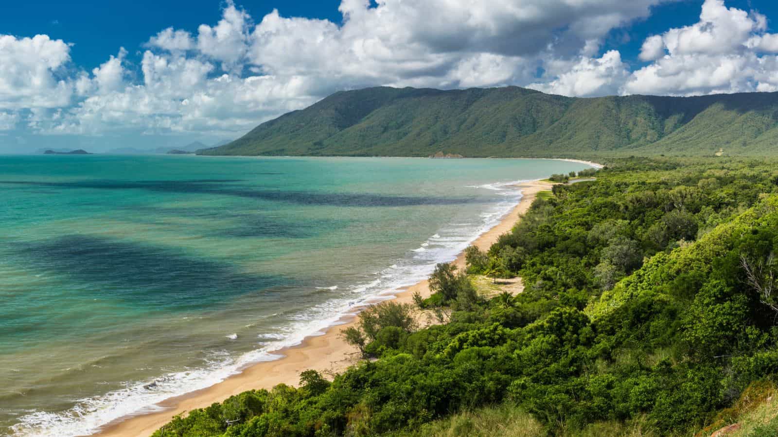 Turquoise water meets a sandy beach, lush forest, and distant mountains beneath a partly cloudy sky.