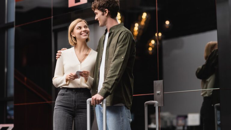 A woman with travel documents and a man with a suitcase smile at each other outside a modern building.