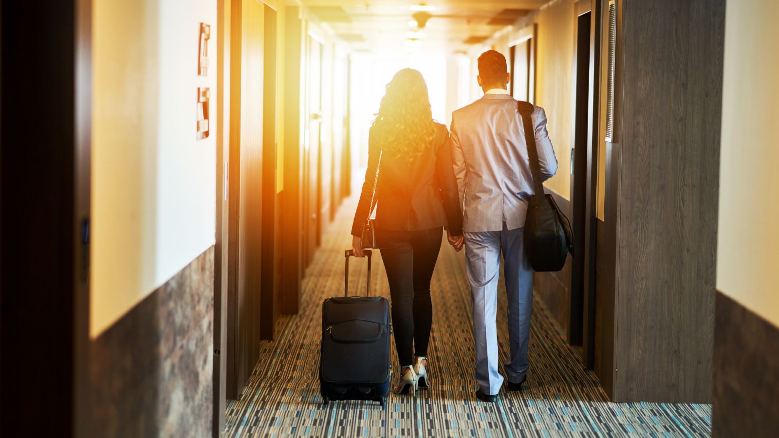 Two people in business attire walk down a hotel hallway with suitcases toward a bright exit.