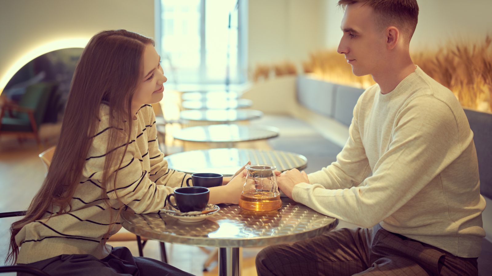 A man and woman sit across from each other at a café table, holding hands, with two mugs and a glass pot between them.