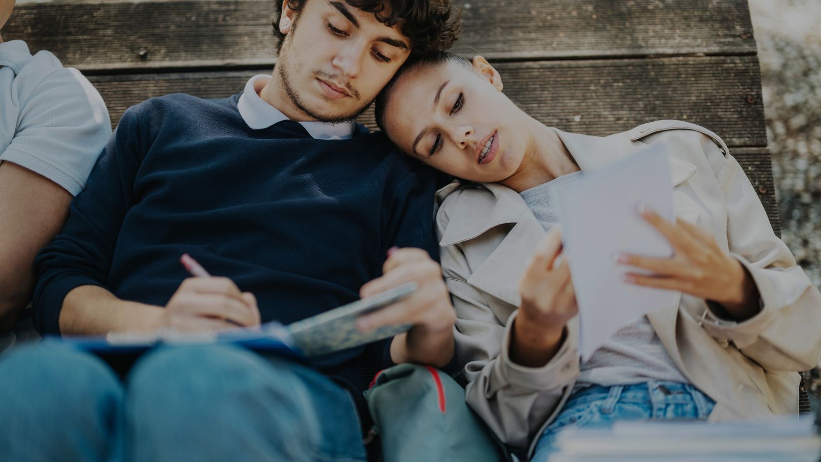 Two people sit close, one writing in a notebook and the other reading a paper, both leaning on each other for support.