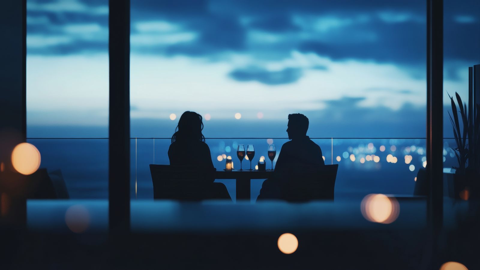 Two people sit at a balcony table with wine glasses at night, city lights blurred in the background.