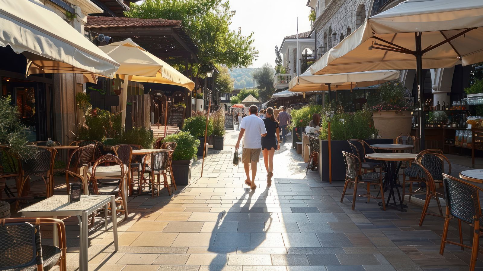 People walk along a sunny pedestrian street lined with outdoor cafés, empty tables, and large umbrellas.
