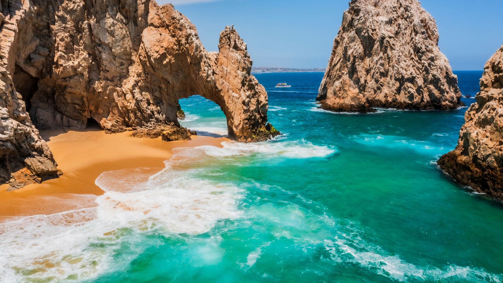 A photo of a dramatic desert cliffs and rock formations meeting deep blue ocean, El Arco visible in the distance, clear sky, rugged natural beauty.
