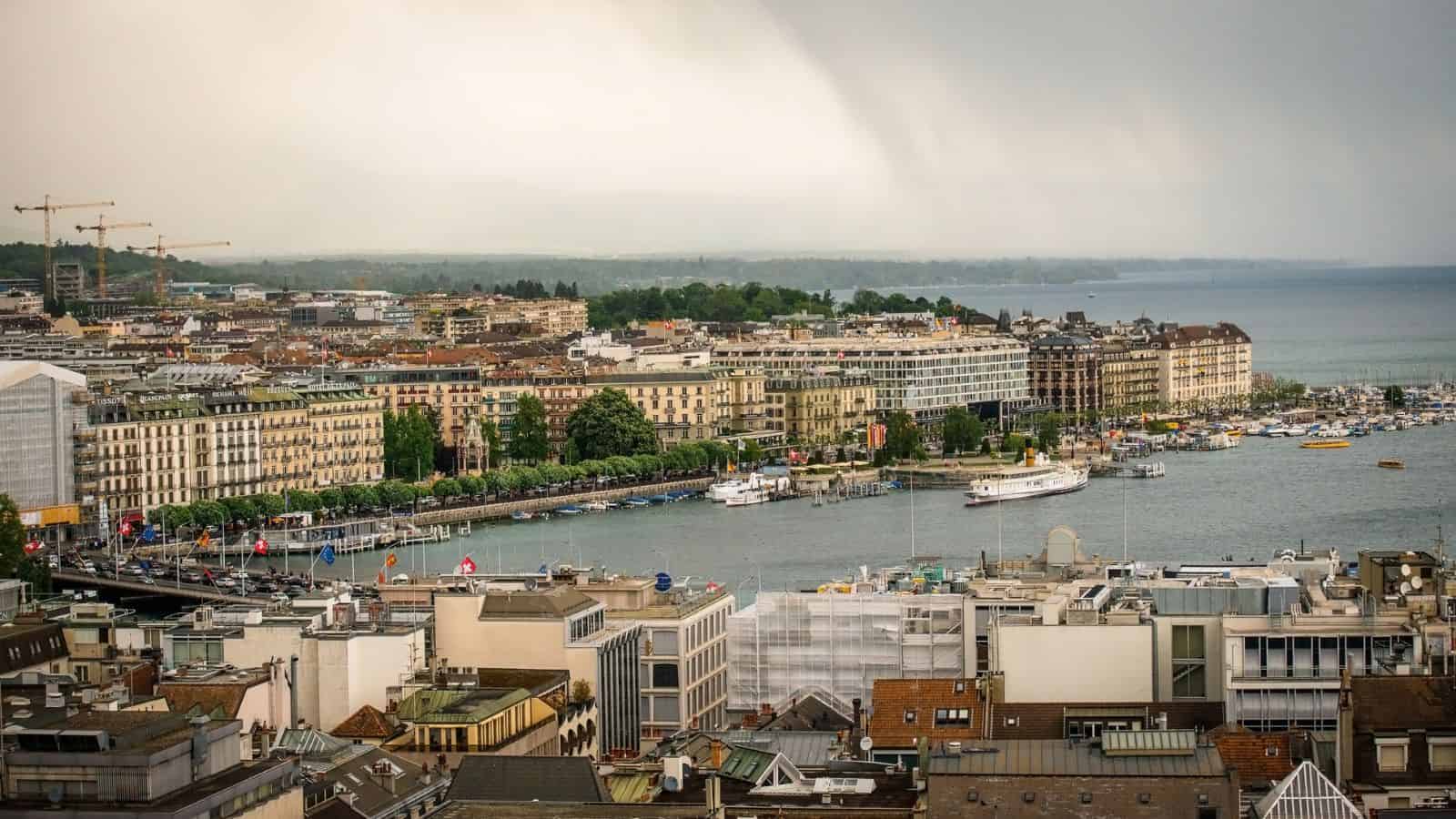 Aerial view of a cityscape featuring a waterfront lined with boats and buildings. The skyline includes construction cranes and a mix of modern and traditional architecture. The sky is overcast, with hints of rain in the background.
