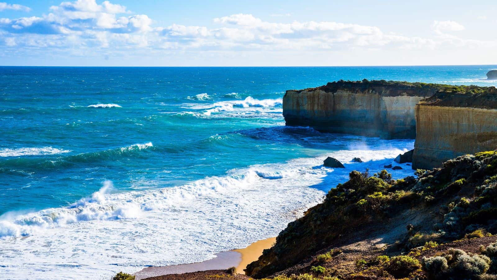 Rocky coastal cliffs rise above blue ocean waves under a partly cloudy sky. The waves crash against the shore, and green shrubs cover the top of the cliffs in the foreground.