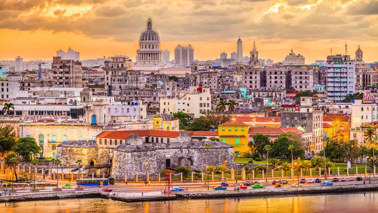 Havana cityscape at sunset with colorful buildings, the Capitolio dome, and historic stone structures by the waterfront.