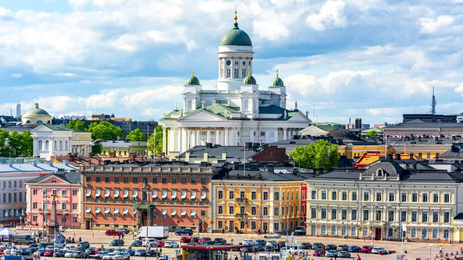 A view of Helsinki Cathedral with its green dome and white facade, surrounded by colorful historic buildings and parked cars under a partly cloudy sky.
