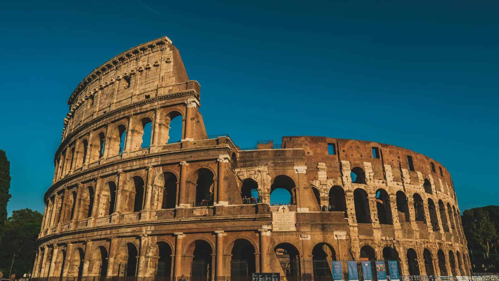The Colosseum in Rome stands beneath a clear blue sky, displaying its ancient arches and partially ruined stone structure.