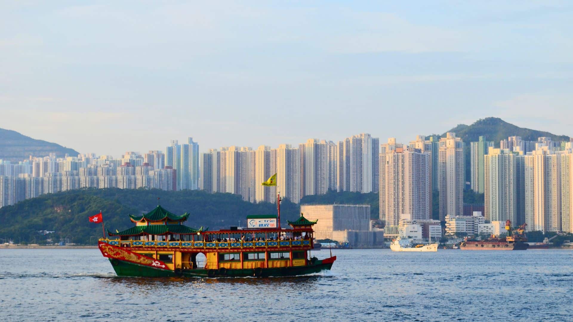 Traditional Chinese boat with ornate decorations sails on water; high-rise buildings and hills in the background.