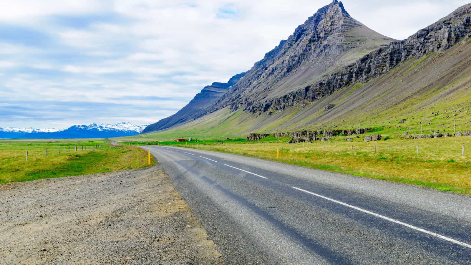 A paved road curves past grassy fields and steep mountains under a mostly cloudy sky.