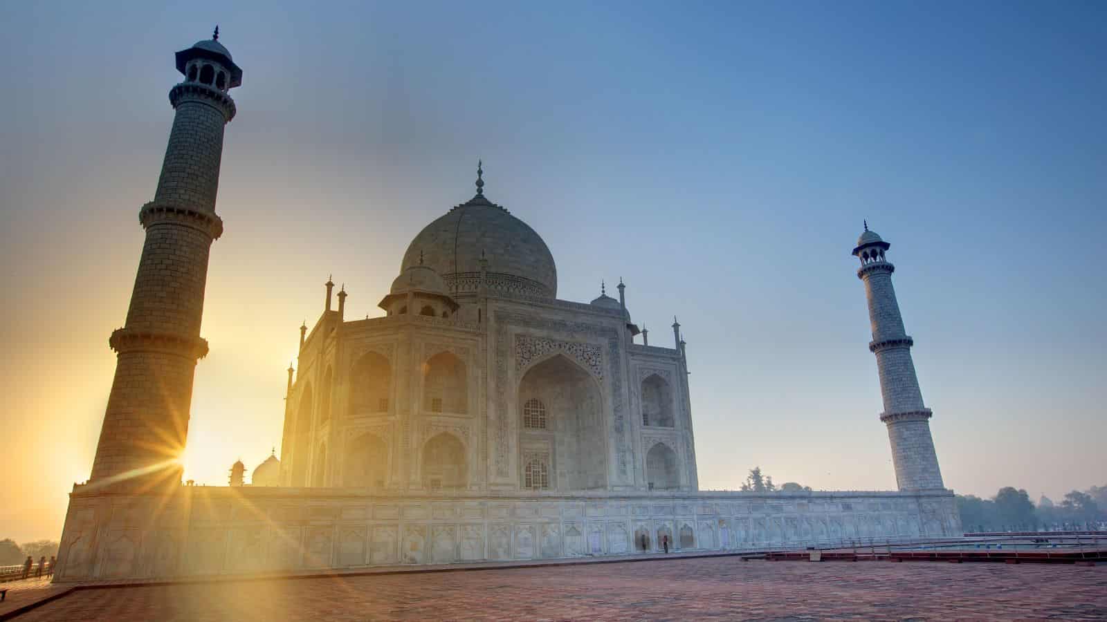 The Taj Mahal at sunrise in Agra, India, its marble dome and two flanking minarets silhouetted against the sky.