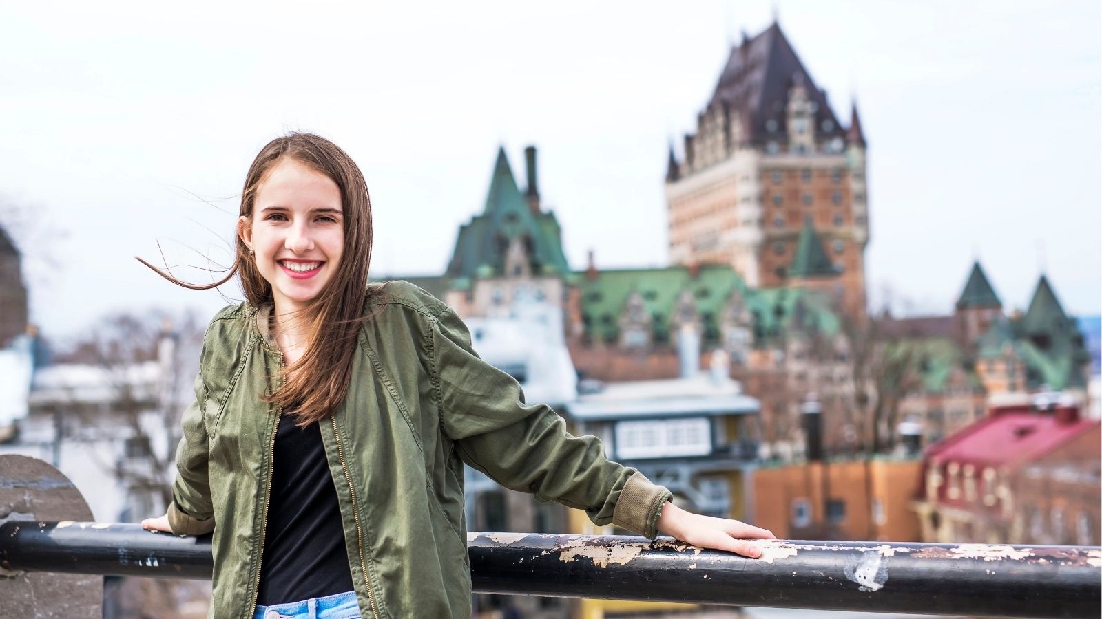 Smiling young woman in a green jacket leans on a railing before historic buildings and a large chateau.