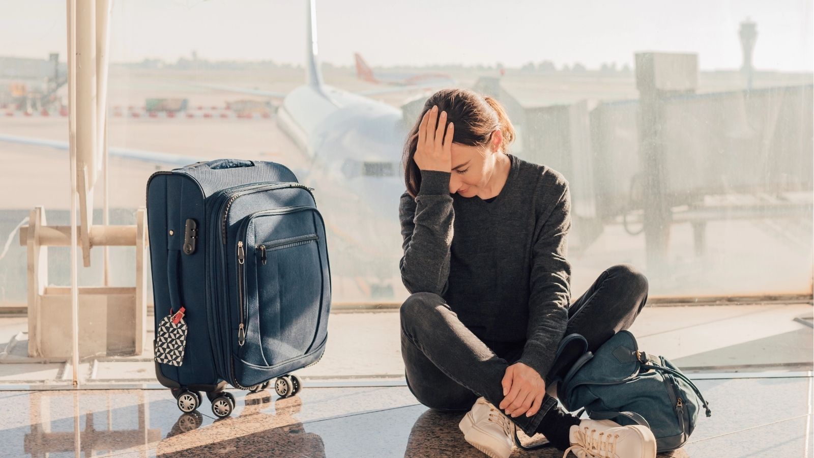 A distressed woman sits on the airport floor with luggage beside her and an airplane visible through the window.