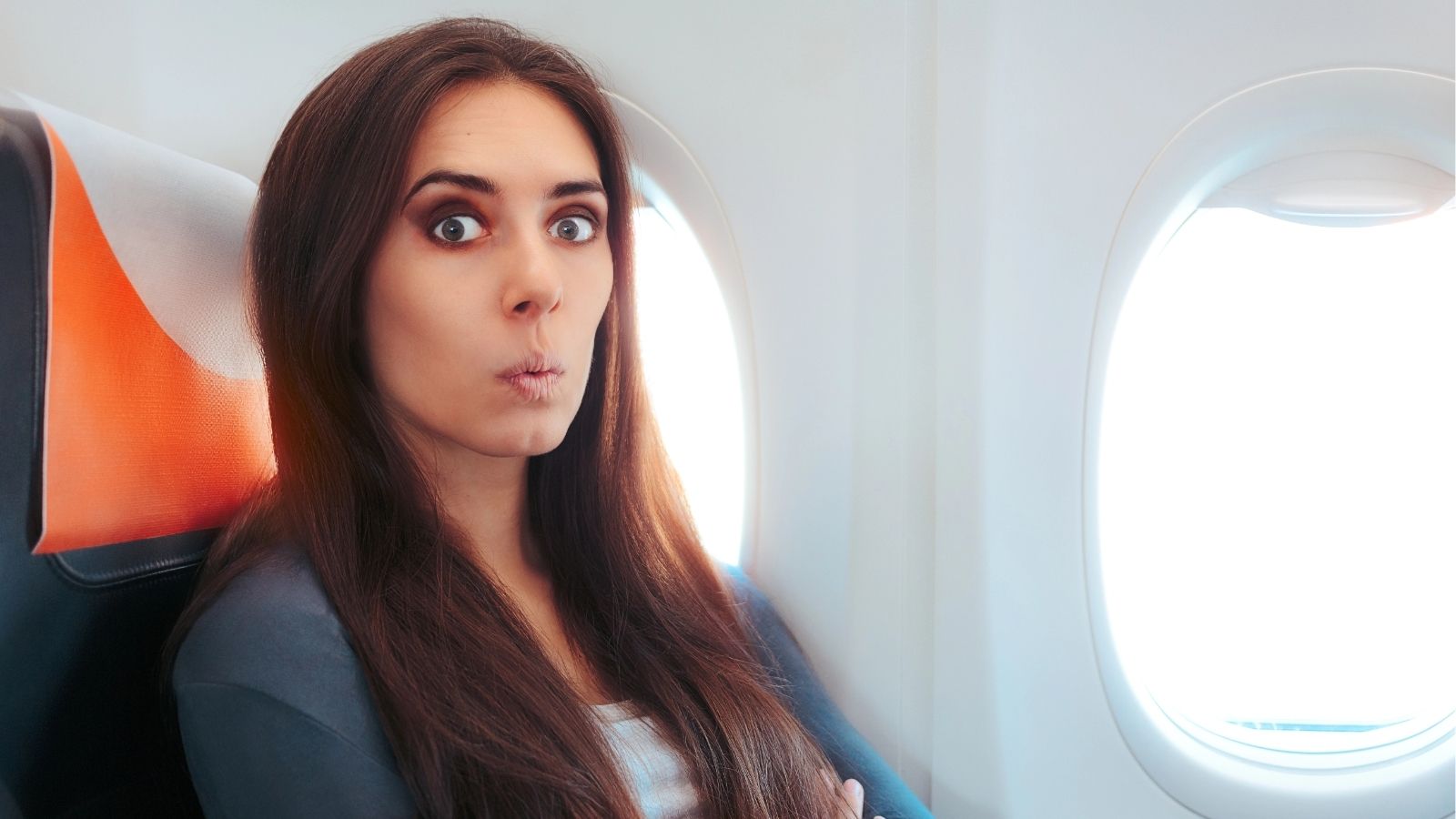 A woman with long brown hair sits by an airplane window, looking surprised with wide eyes and puckered lips.