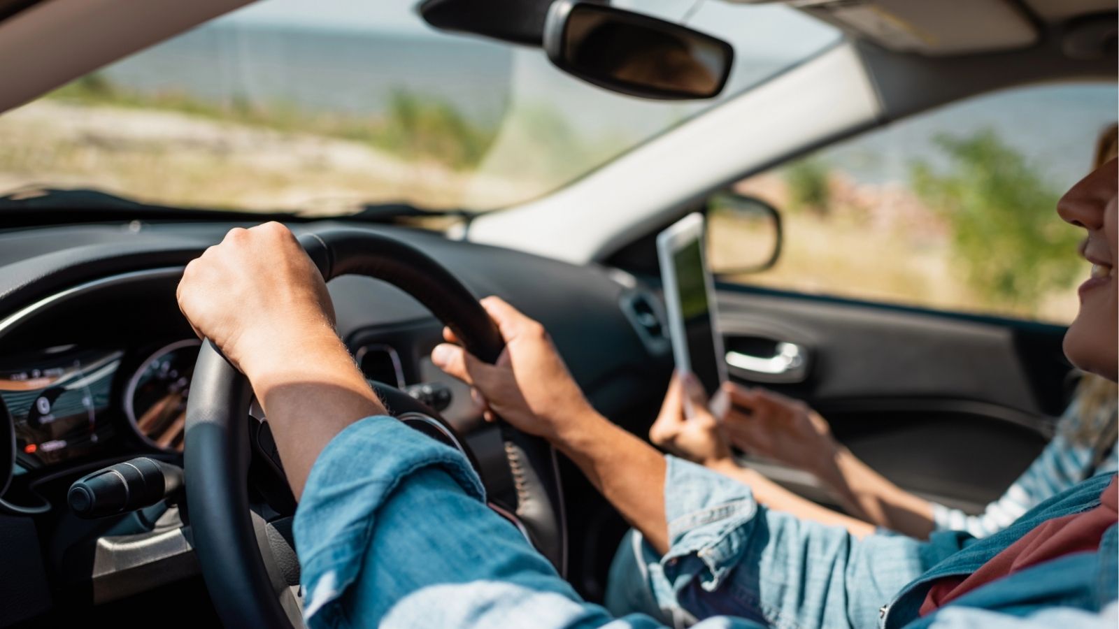 Two people sit in a brightly lit car; the driver holds the wheel while the passenger uses a tablet against a blurred background.