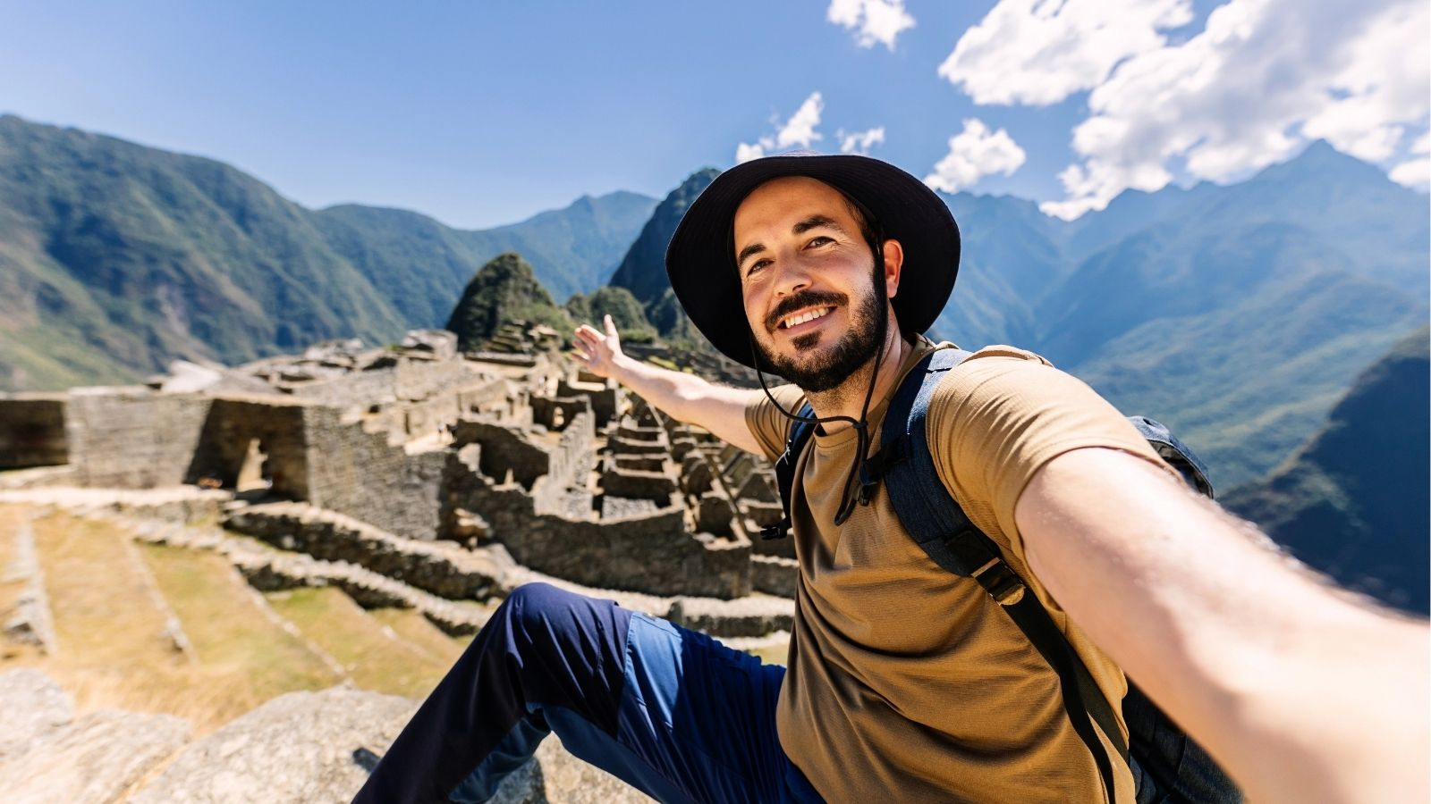 Man in hat and backpack smiles for a selfie, pointing at Machu Picchu’s stone ruins with mountains behind under clear sky.