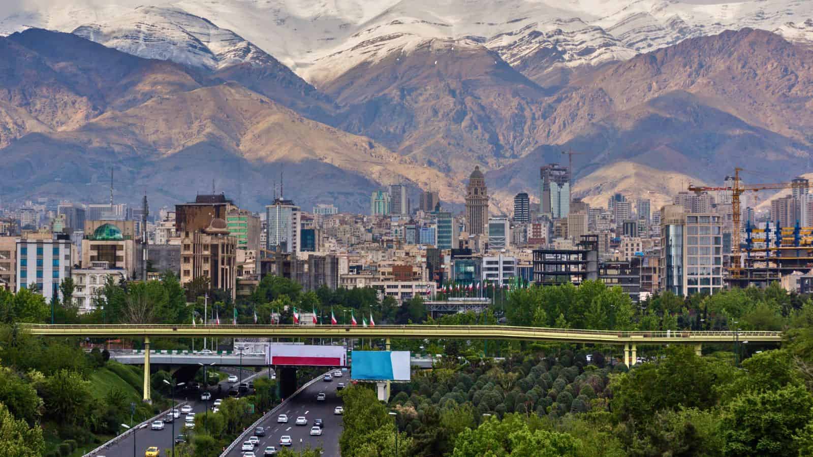 Modern city buildings, a car-filled highway, green trees, and snow-capped mountains in the background.