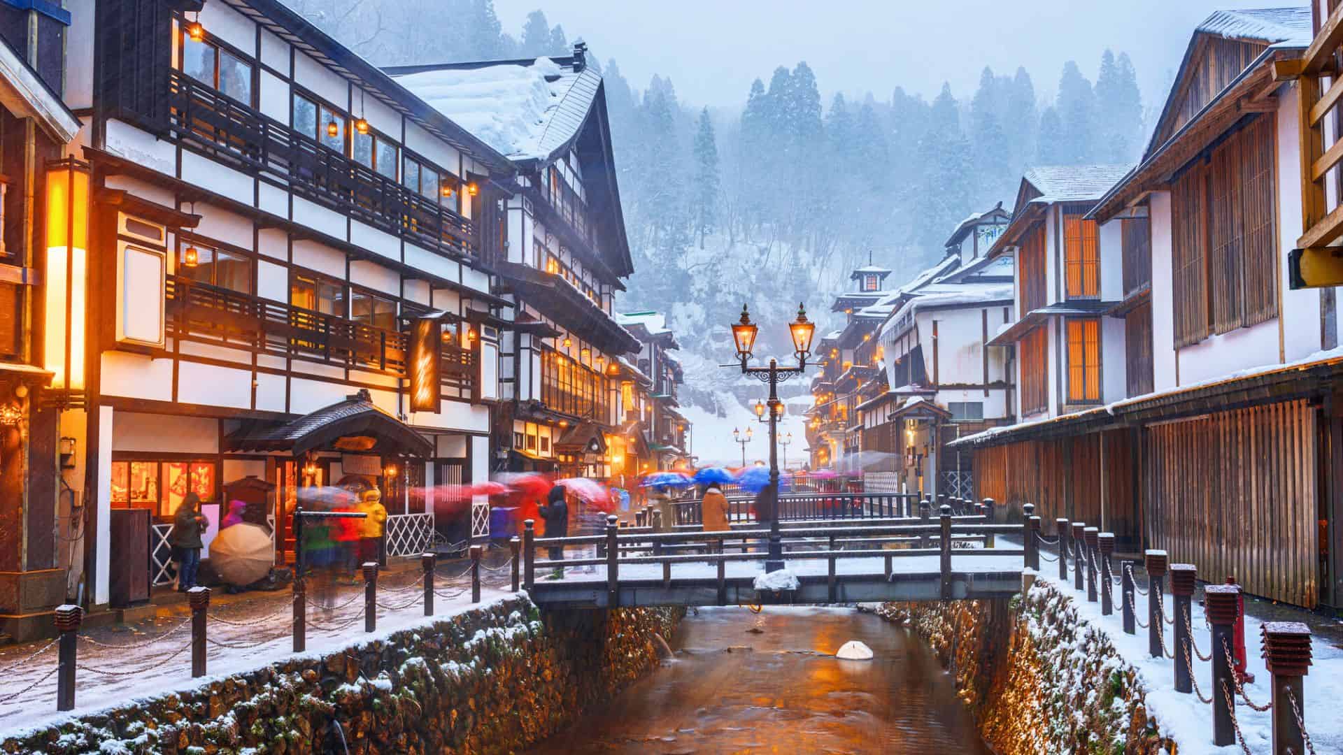Snow falls on a Japanese village with wooden houses, a lit bridge over a stream, and people carrying umbrellas.