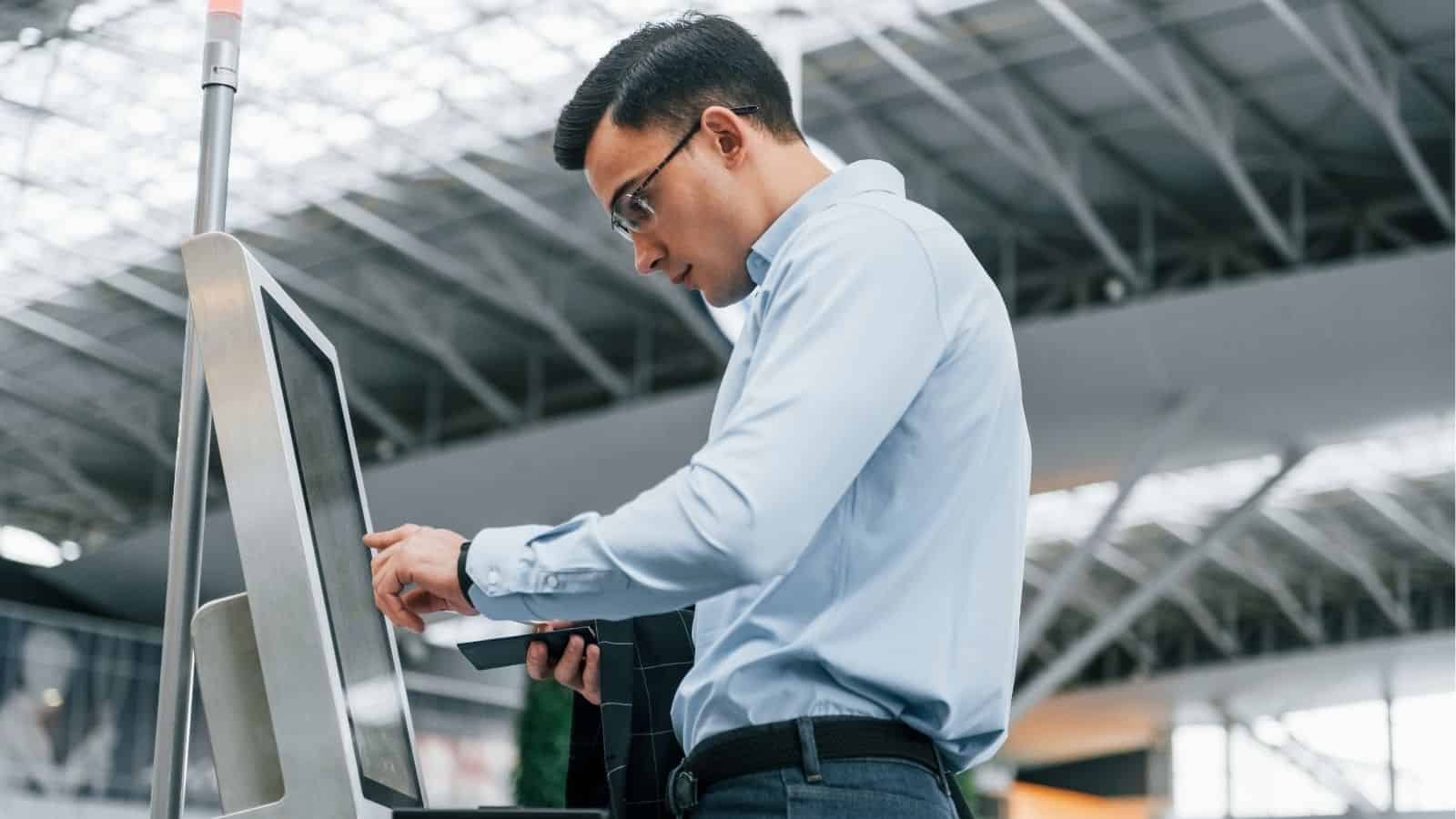 A man in a light blue shirt uses a self-service kiosk in an indoor public space, likely an airport or station.