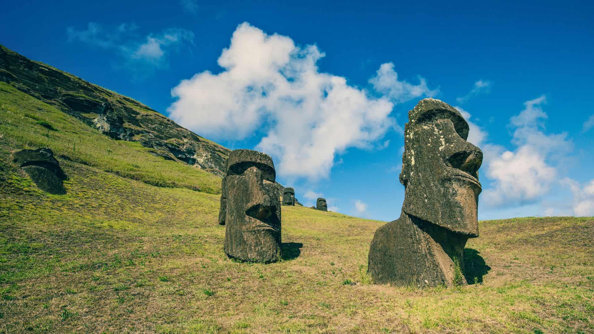 Two stone Moai statues stand on a grassy hill beneath a blue sky with scattered clouds on Easter Island.