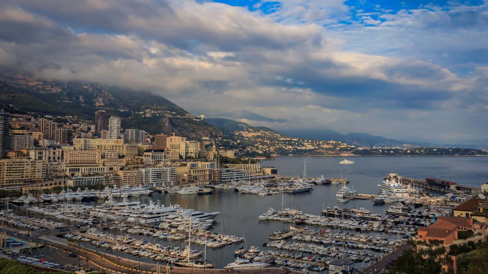 A harbor filled with numerous yachts and boats is surrounded by tall buildings and hills under a mostly cloudy sky, with mountains in the background.