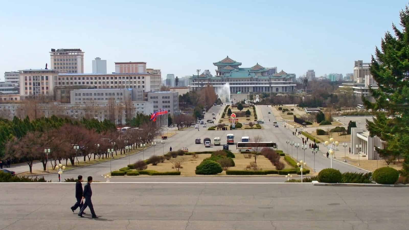 City avenue with pedestrians, traffic, and a large traditional-style building at the end amid trees and modern buildings.