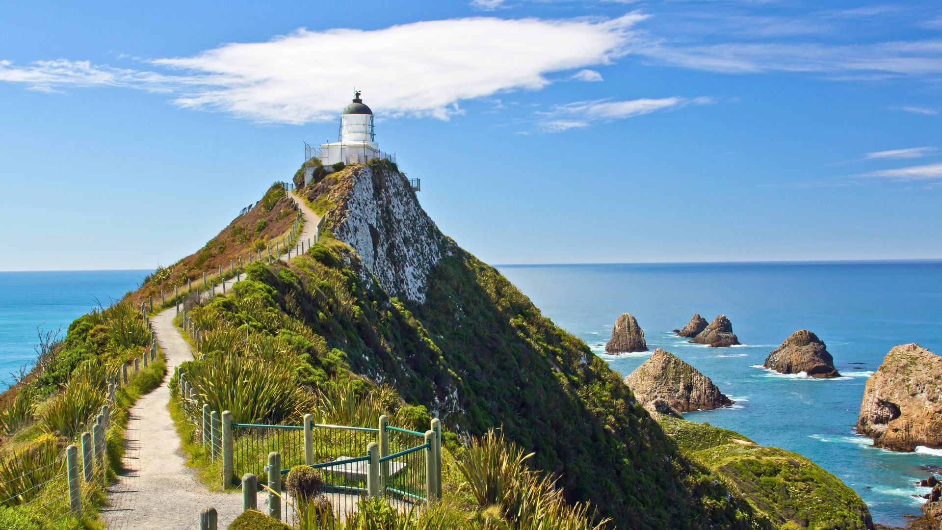A lighthouse stands on a grassy hill above the ocean, with rocky islets nearby and a clear blue sky overhead.